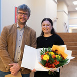 Dr. Najla Sadat und Dr. Daniel Wiswede stehen nebeneinander und schauen in die Kamera. Sadat hält eine Urkunde und einen Blumenstrauß.