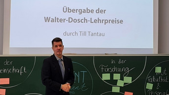 Eine Person steht vor einer Tafel. Über der Tafel wird eine Präsentation angeezeigt, auf der steht: Übergabe der Walter-Dosch-Lehrpreise durch Till Tantau.